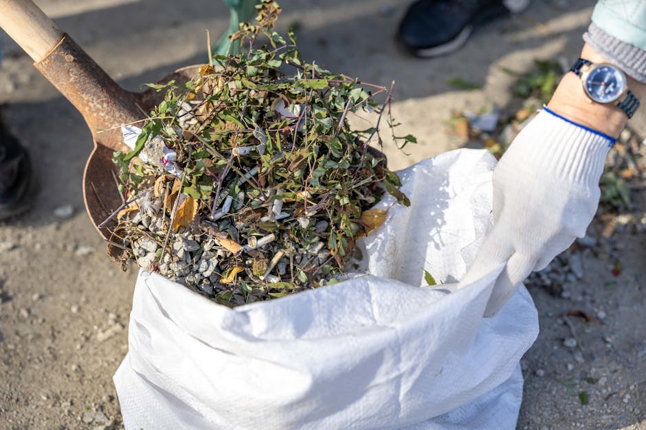 A person wearing a white glove and a wristwatch is using a shovel to scoop up a mixture of small rocks, dry leaves, and trimmed plant branches into a large white waste bag outside on a gravel surface.