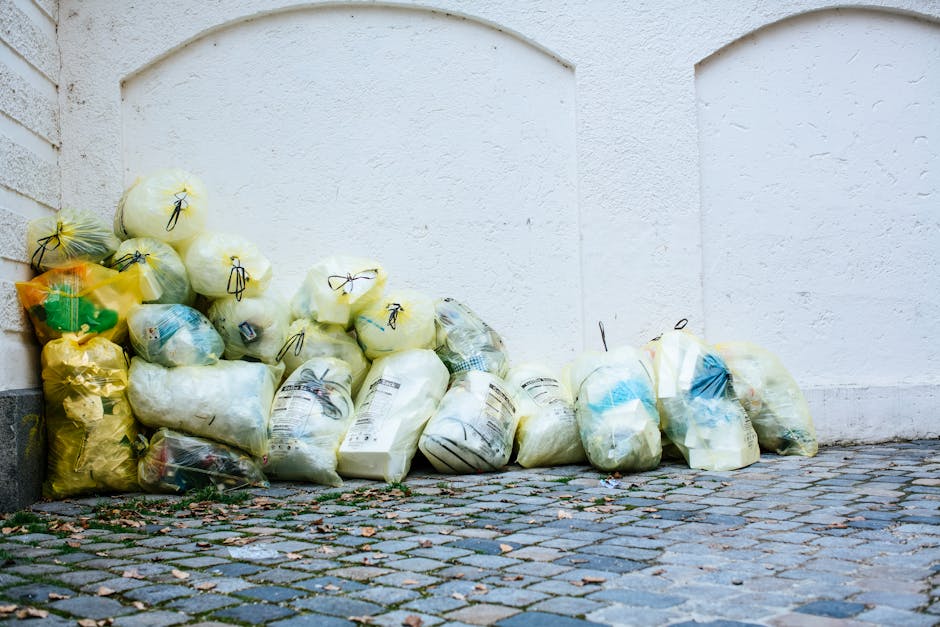 A person wearing a white glove and a wristwatch is using a shovel to scoop up a mixture of small rocks, dry leaves, and trimmed plant branches into a large white waste bag outside on a gravel surface.