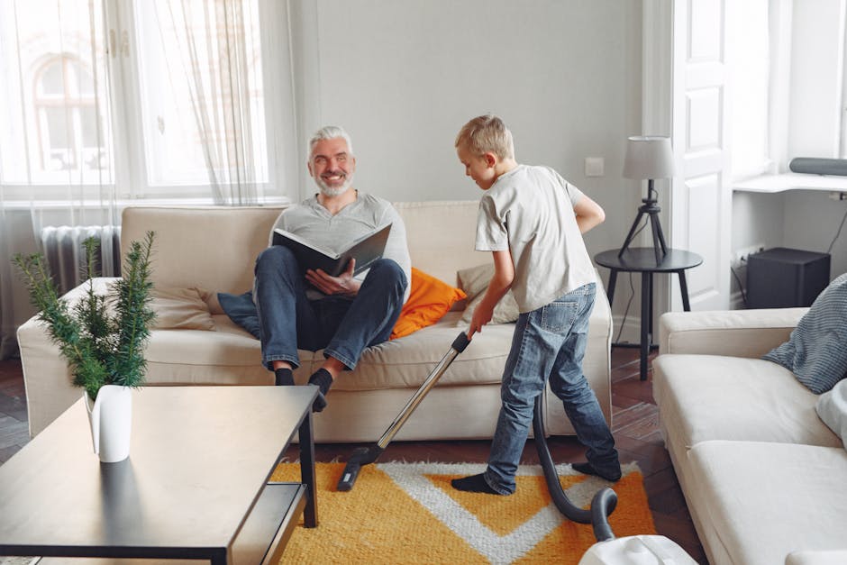 A young boy vacuuming a beige fabric sofa in a bright, modern living room with natural light coming through white-framed windows. The boy is dressed in a light gray T-shirt and blue jeans, focused on 