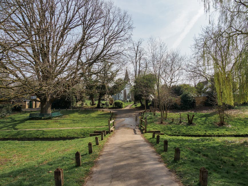A paved pathway in a park during late winter or early spring, with leafless trees on either side and some budding greenery. The path is bordered by small wooden posts and leads towards a small stone b